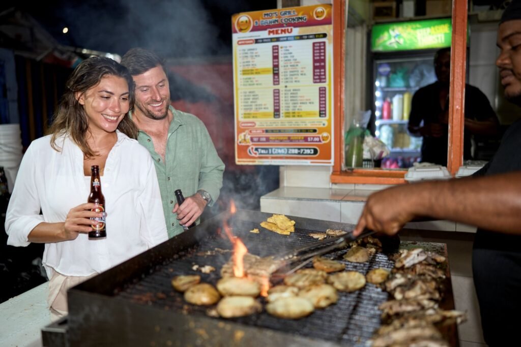 Pareja disfrutando la gastronomía local en Barbados, con parrillas de pescado fresco y cerveza caribeña.