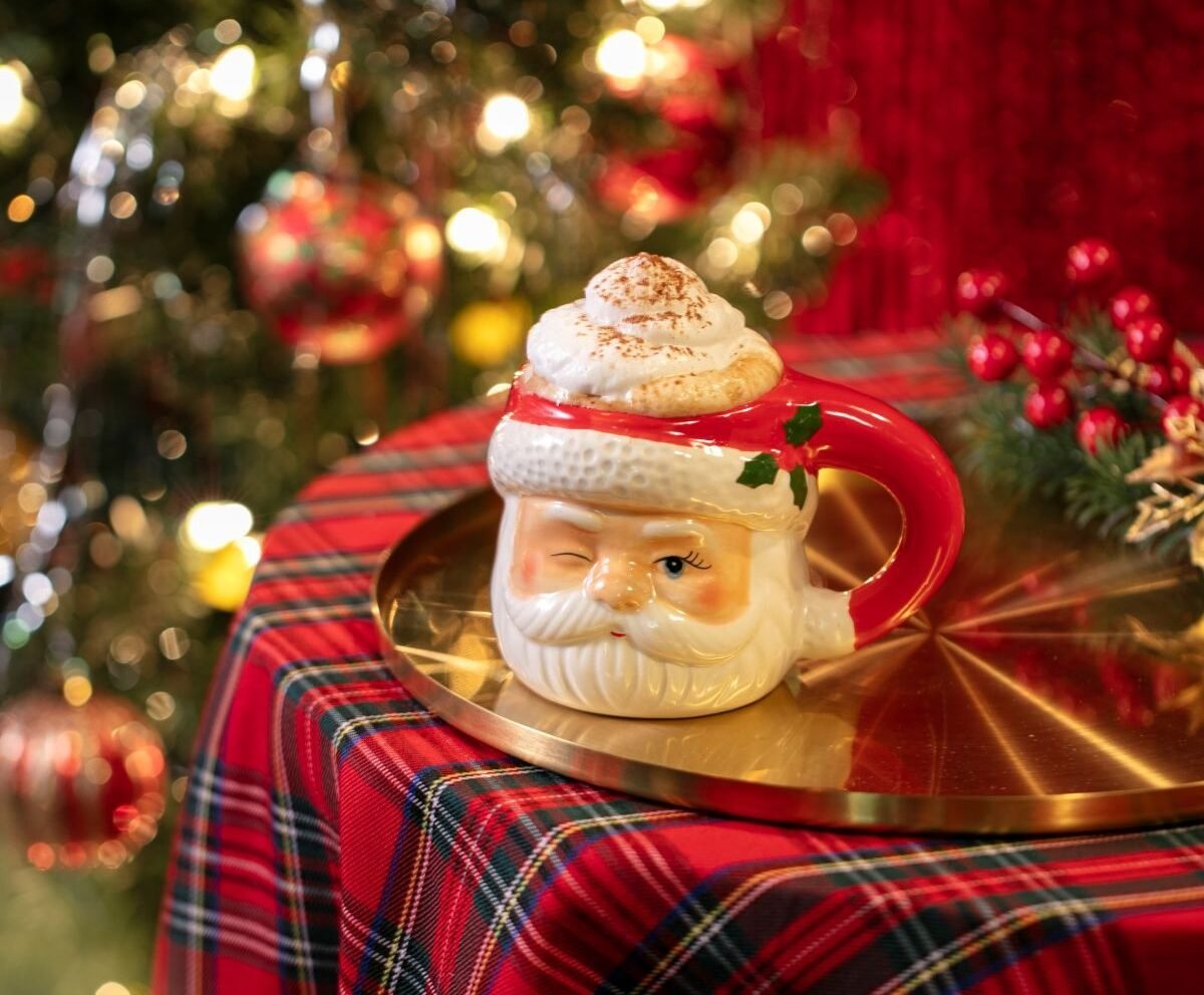 Taza navideña con forma de Santa Claus llena de chocolate caliente con crema batida, frente a un árbol de Navidad iluminado.